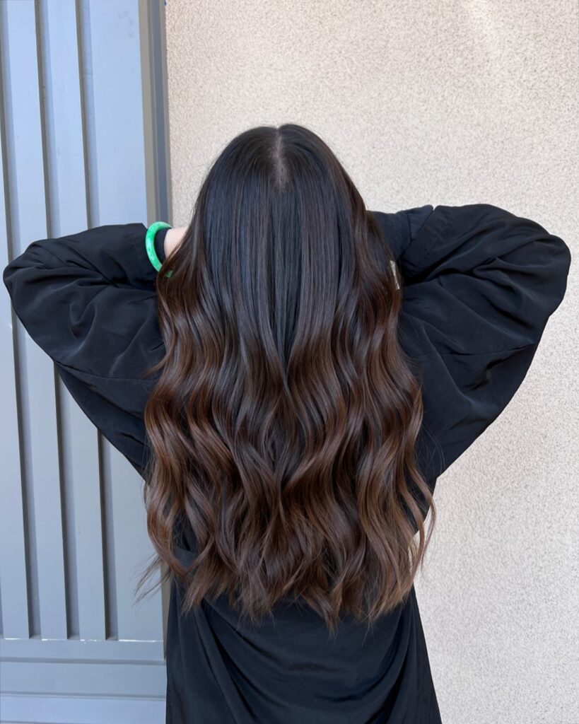 A back view of a woman with long dark brown hair styled in glossy waves with subtle chocolate balayage ends.