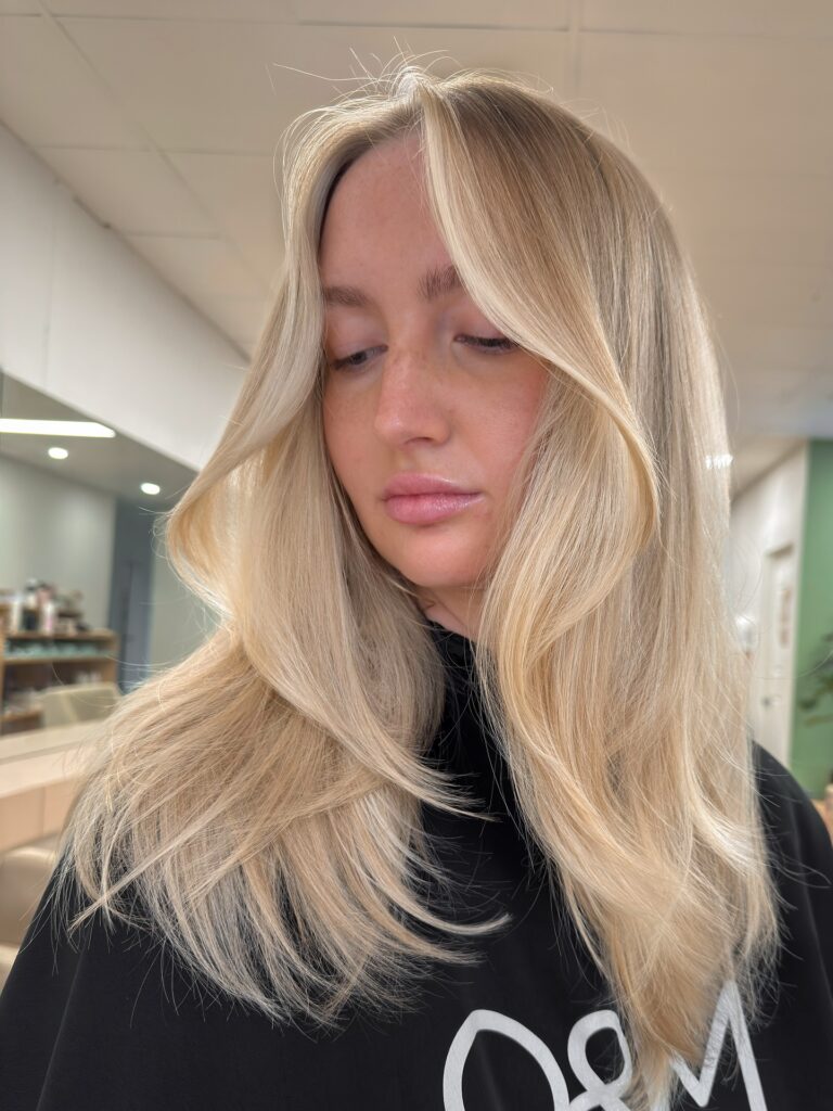 A woman with long layered blonde hair and face framing highlights styled in a voluminous blowout.