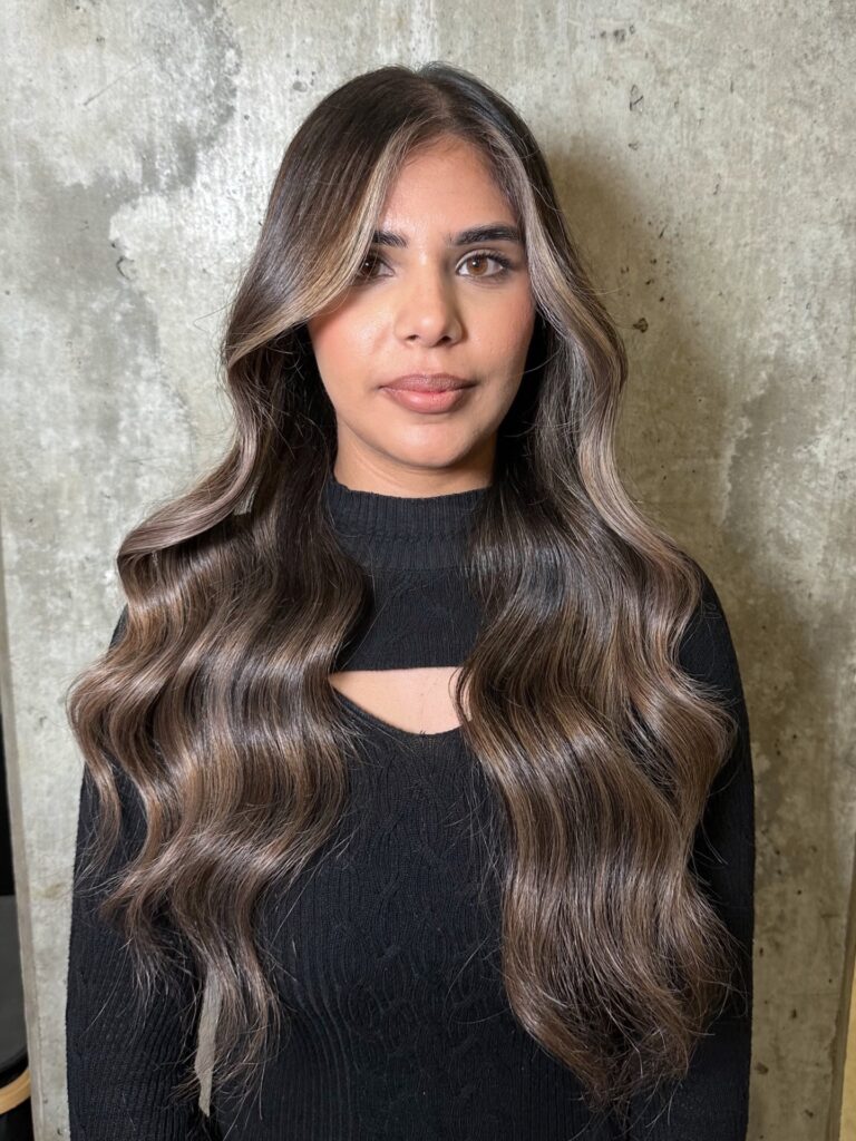 A woman with very long dark brown hair styled in structured waves with bright blonde face framing highlights.