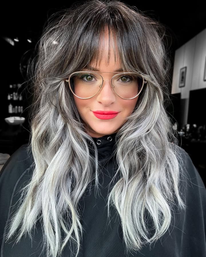 Long wavy gray ombre hair with full textured bangs and dark roots.