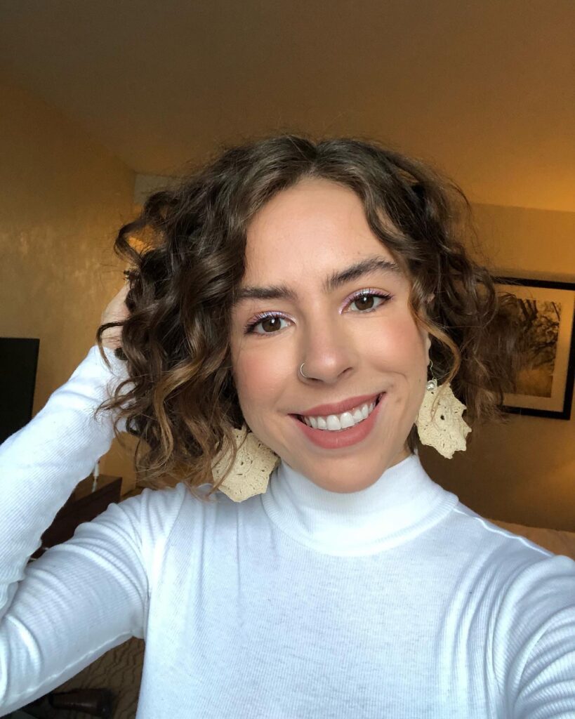 A smiling woman with a chin-length curly brunette bob featuring subtle blonde highlights and a middle part.