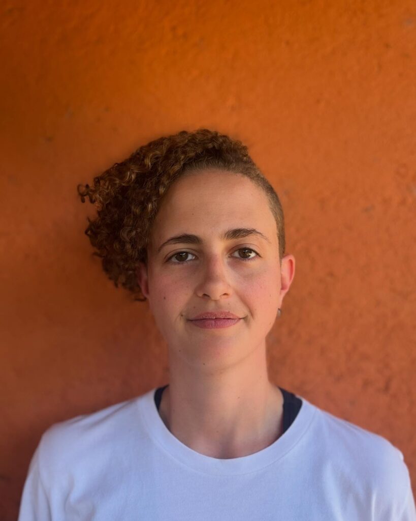 A person with an asymmetrical curly hairstyle featuring a side shave and voluminous honey brown ringlets swept to one side.
