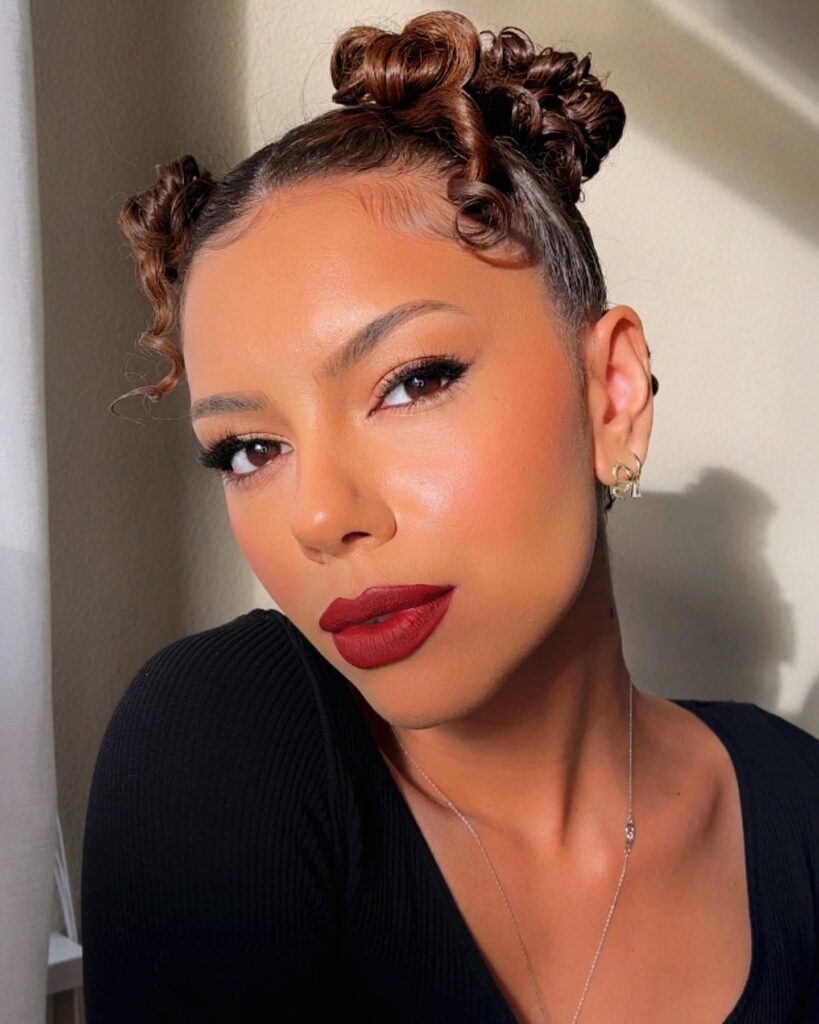 A woman with mocha brown hair styled into two curly space buns with sleek edges and a few loose ringlets.