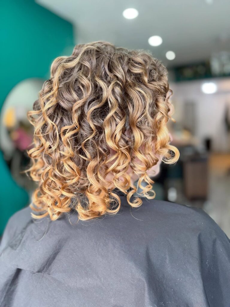 A back view of a woman with a shoulder-length curly bob featuring honey blonde highlights and defined ringlets.