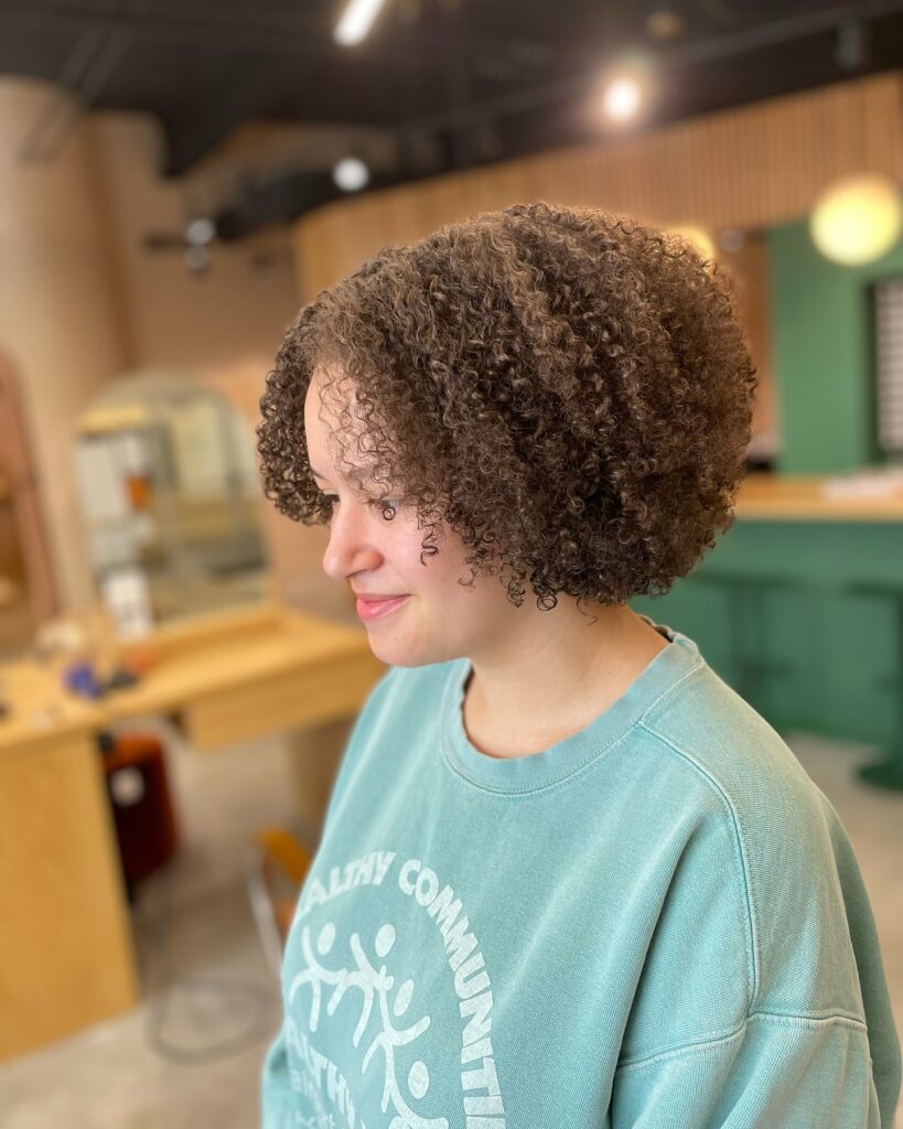 A woman with a rounded brunette afro featuring soft, defined natural ringlets and a voluminous halo shape.