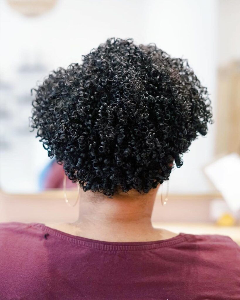 A back view of a woman with a short, heart-shaped hairstyle featuring highly defined natural black coils.