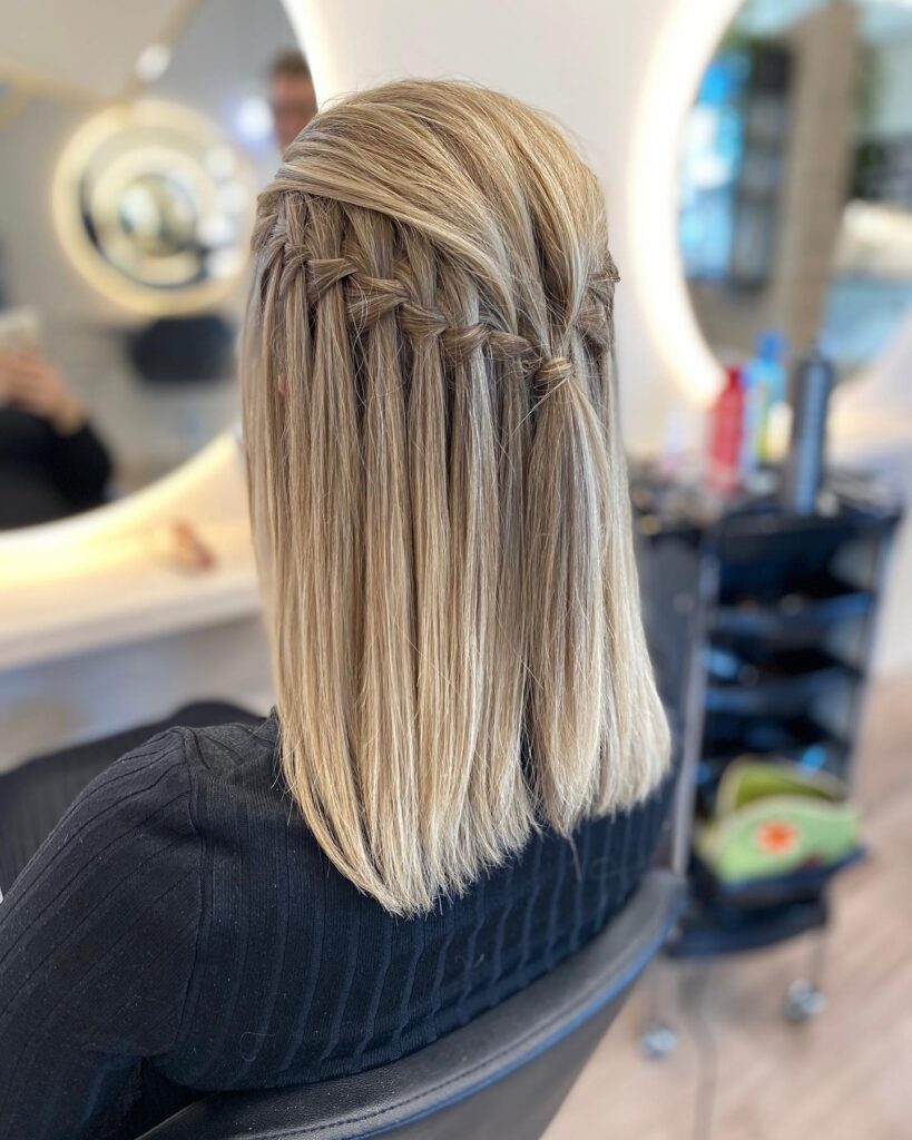 A back view of a woman with straight, sandy blonde, shoulder-length hair featuring a precisely woven waterfall braid.