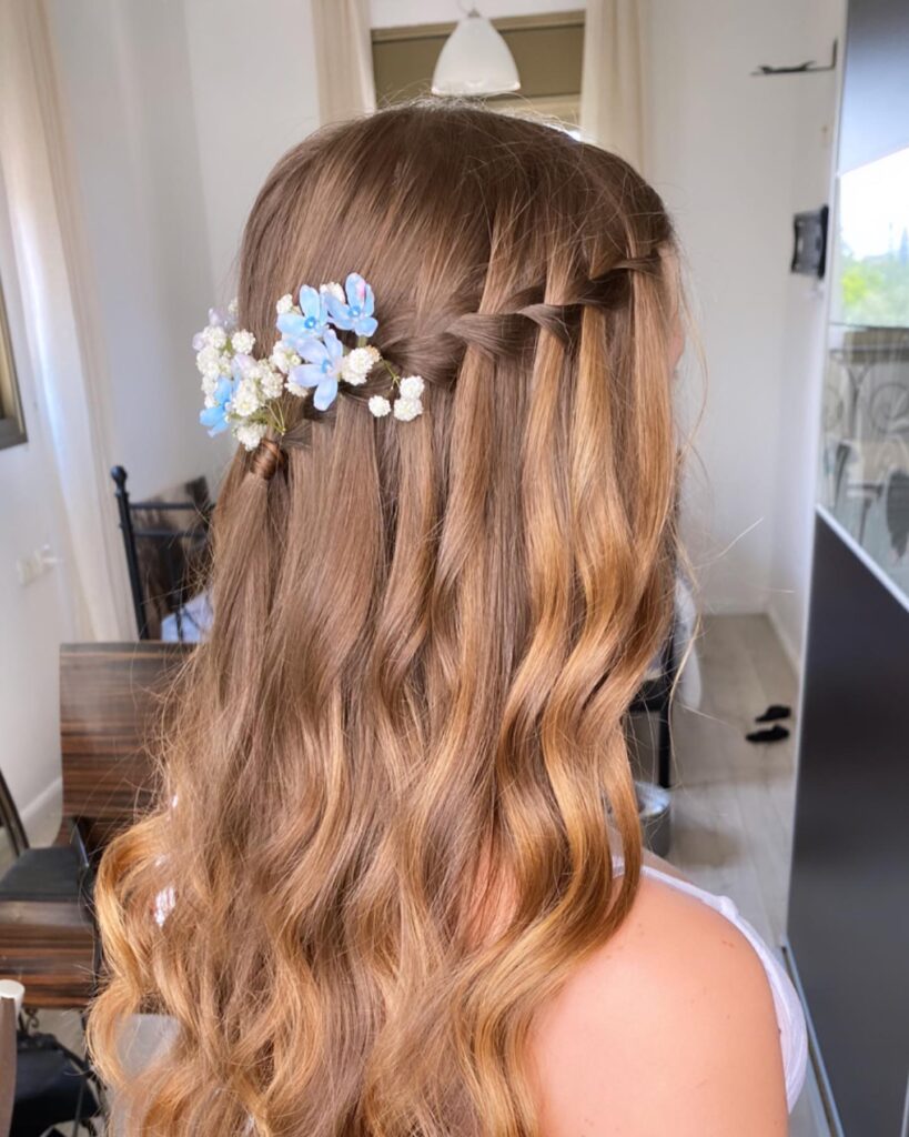 A side view of long, wavy, caramel brown hair featuring a waterfall braid adorned with small blue and white flowers.