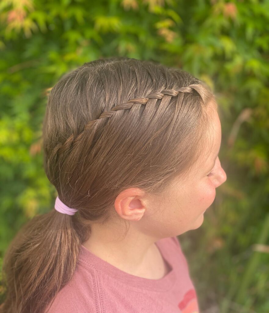 A side profile of a girl with ash brown hair styled in a thin waterfall braid that leads into a low ponytail secured with a pink hair tie.