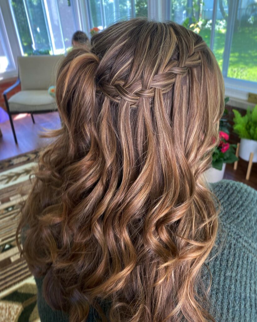 A back view of a woman with highlighted bronde hair styled in a horizontal waterfall braid that flows into thick, bouncy curls.