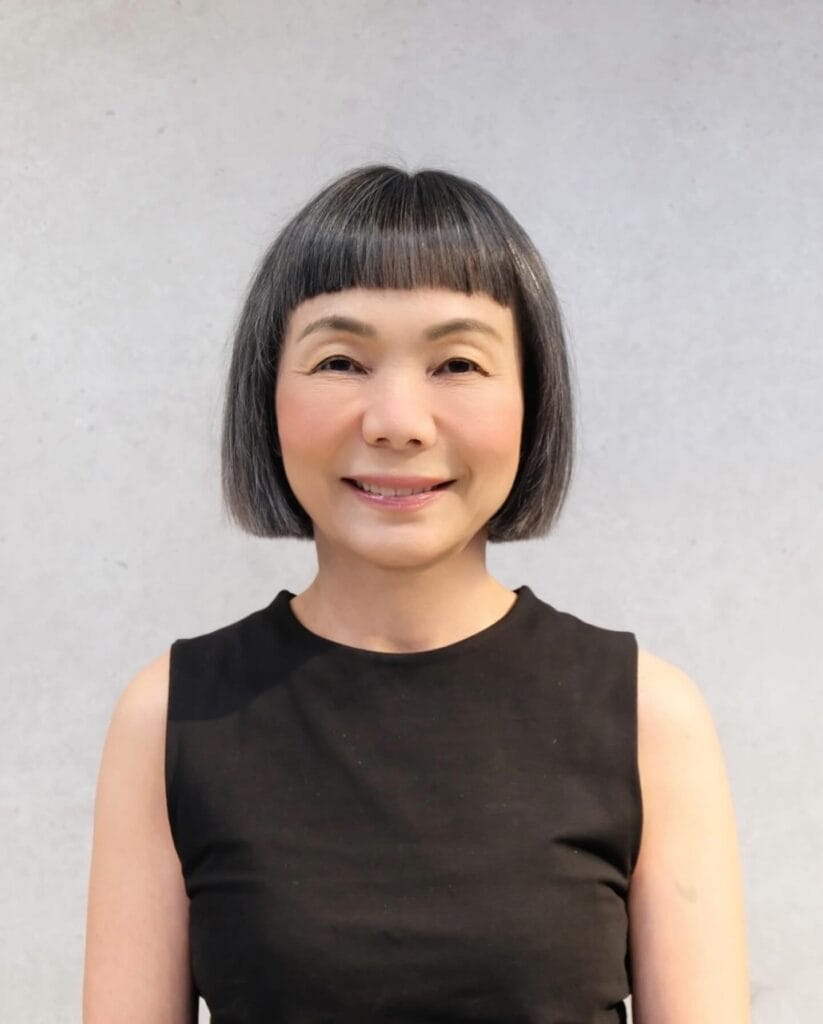 An older Asian woman with a sharp, blunt jaw-length bob haircut and short microbangs in a salt-and-pepper gray color against a light background.