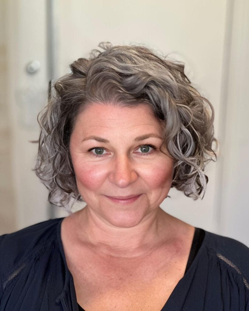 A woman with a chin-length curly bob hairstyle in a mix of gray and pewter tones featuring soft natural layers.