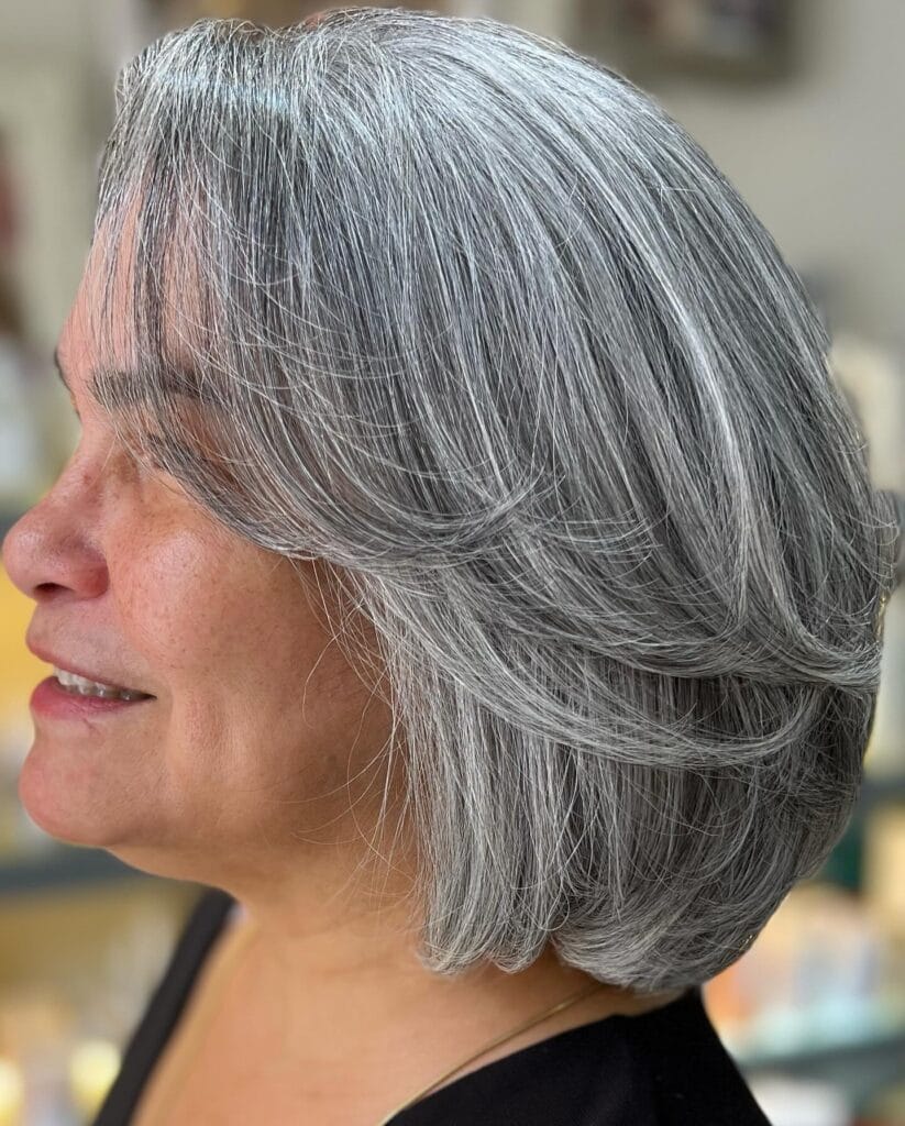 A side profile view of a woman with a voluminous silver-feathered bob hairstyle featuring soft swept-back layers and a rounded back.