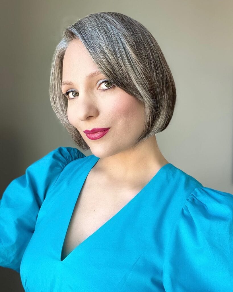 A woman with a sleek chin-length gray bob featuring a deep side part and smooth, curved ends, wearing a bright blue top.