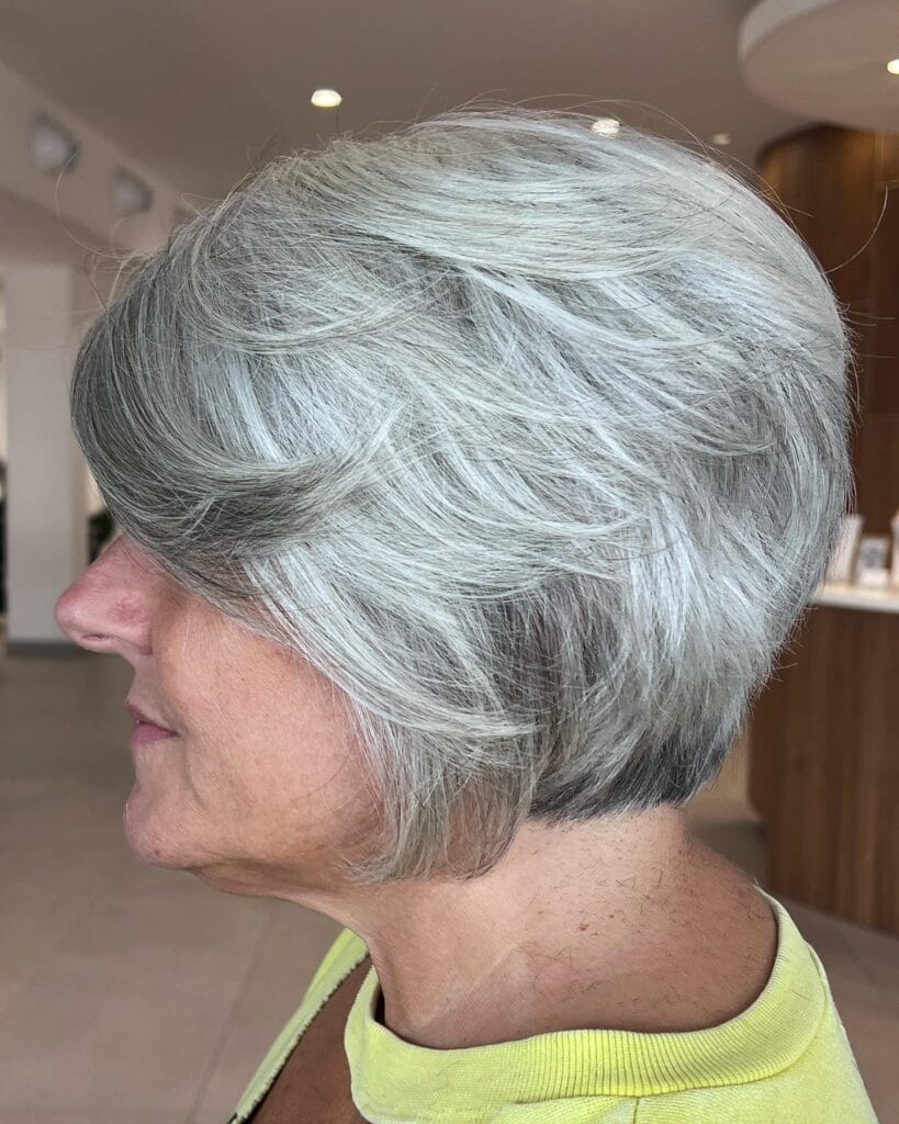 A side profile view of a woman with a layered pewter silver bob hairstyle featuring wispy feathered layers and a graduated back.