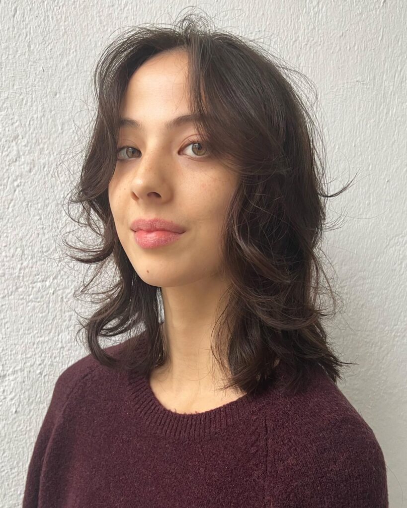 A woman with a medium length dark brown wolf cut featuring shaggy layers and soft curtain bangs.