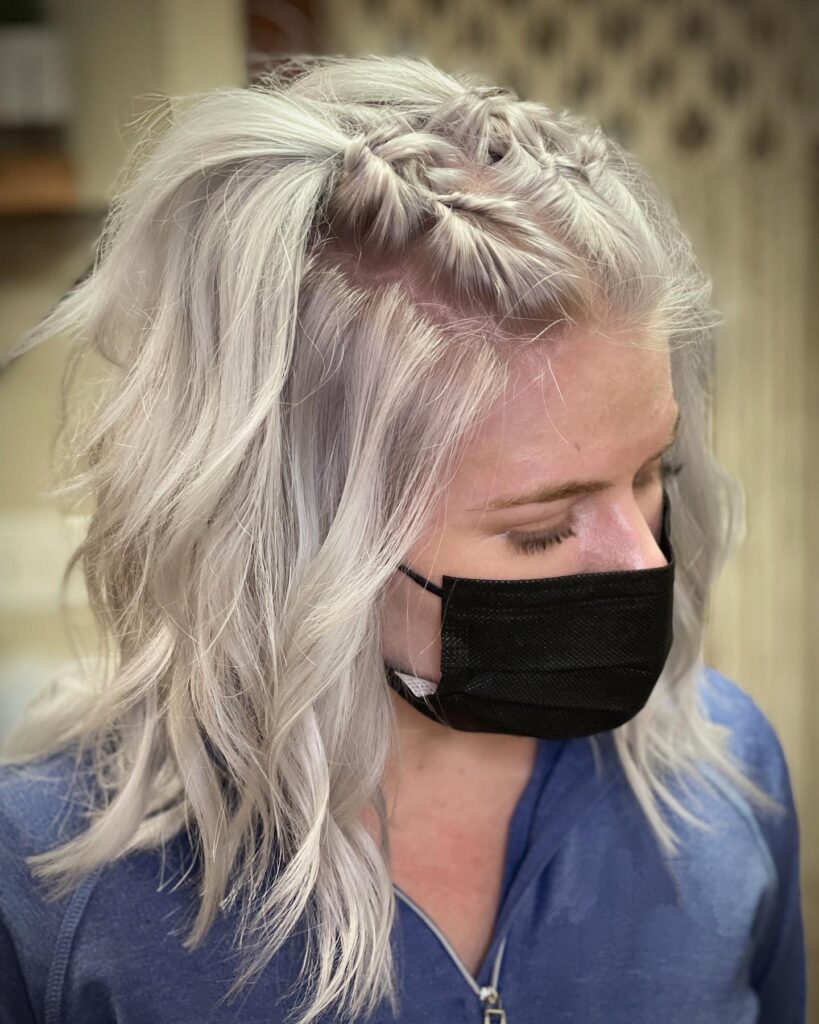 A woman with medium-length icy silver hair styled in loose waves with two small braids on top.