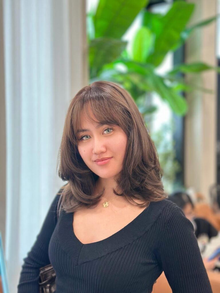 A young woman with medium-length ash brown hair featuring wispy bangs and soft face-framing layers.