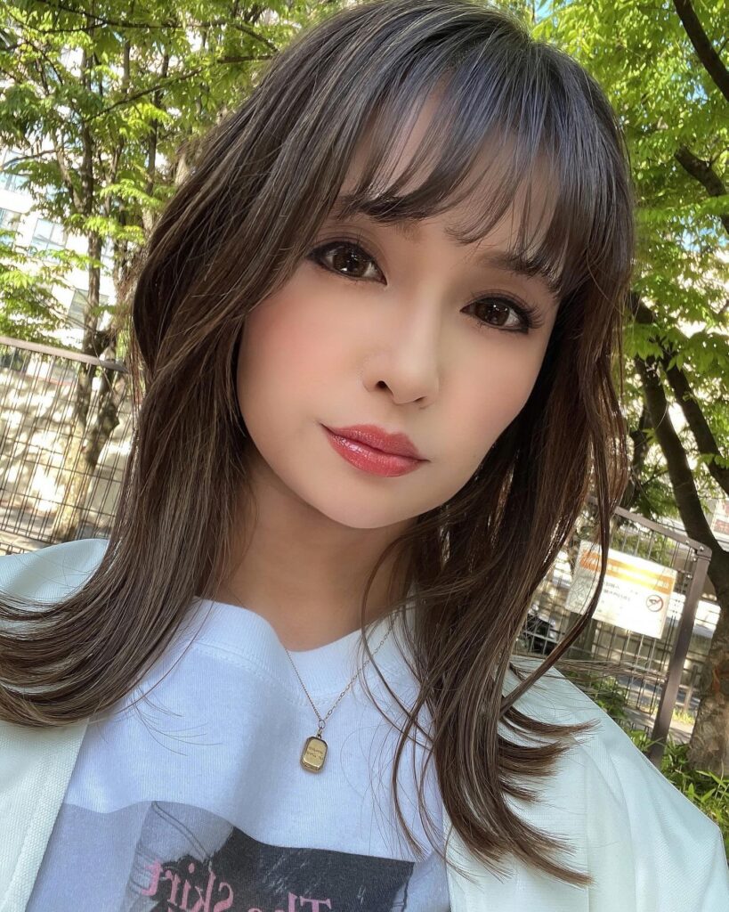 A woman with medium-length, mushroom-brown hair featuring airy, see-through bangs and wispy layers at the ends.
