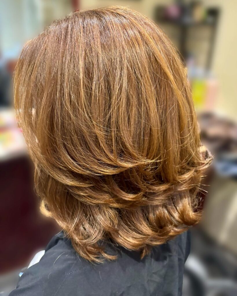 A back view of a woman with shoulder-length warm caramel hair featuring voluminous tiered layers and flipped ends.