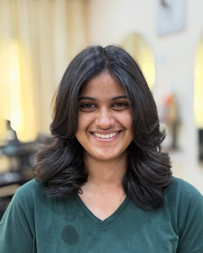 A smiling woman with medium-length dark brunette hair styled with voluminous butterfly layers and large curls at the ends.