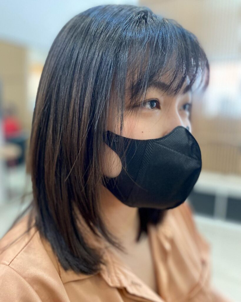 A side profile of a woman with sleek, straight, medium-length, dark hair and thin wispy bangs.