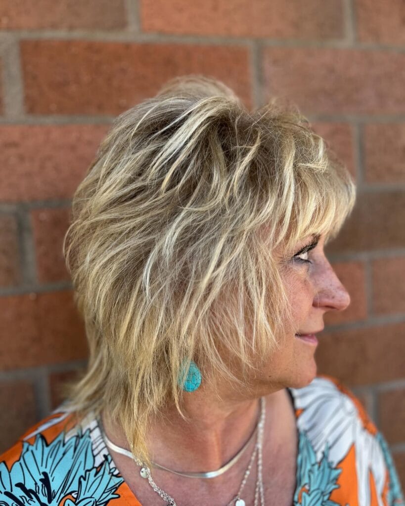 A side profile of a woman with a medium-length golden blonde shaggy haircut featuring voluminous feathered layers and wispy bangs.