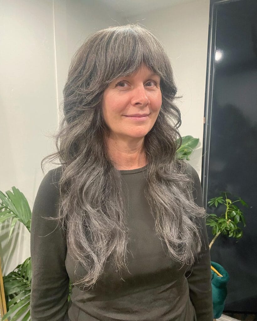 A woman with very long, charcoal and silver shaggy hair featuring voluminous layers and a thick, feathered fringe.