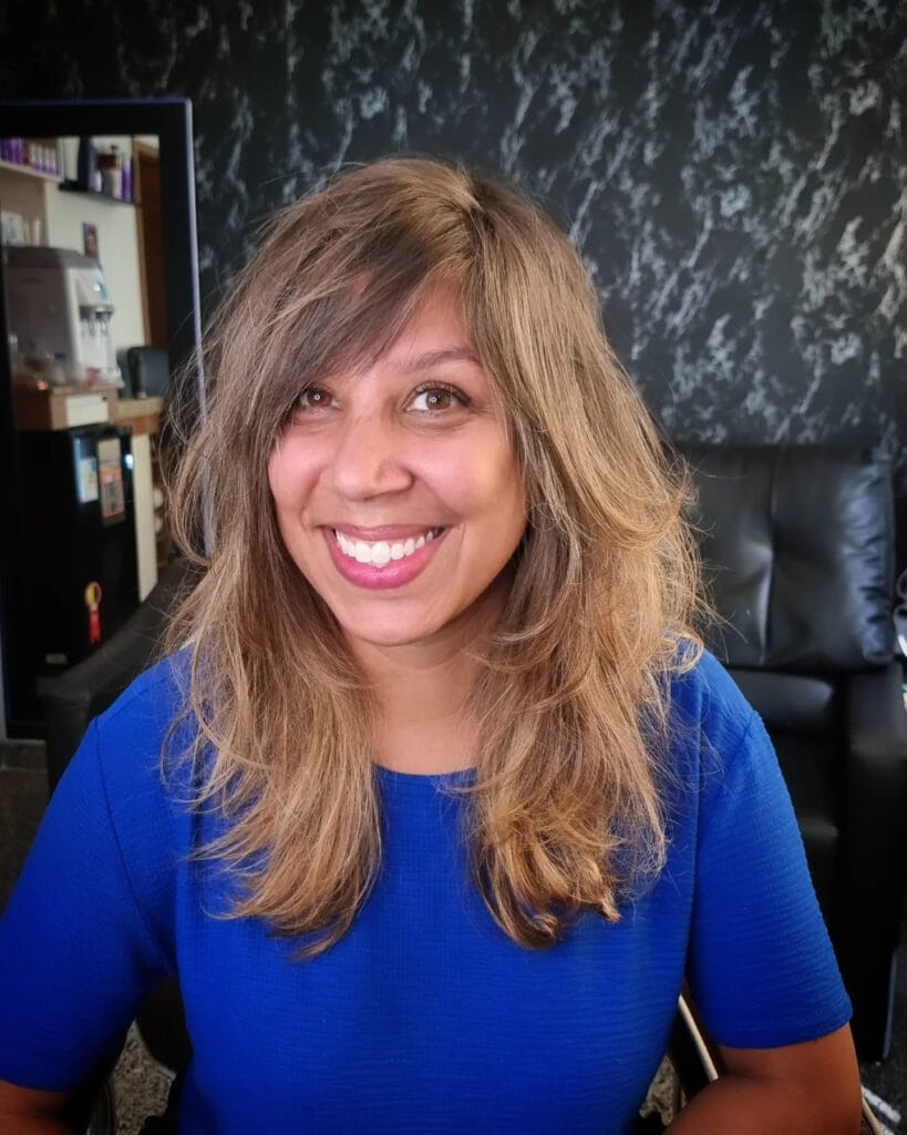 A smiling woman with long, honey brown, shaggy hair featuring a side-swept fringe and textured layers.