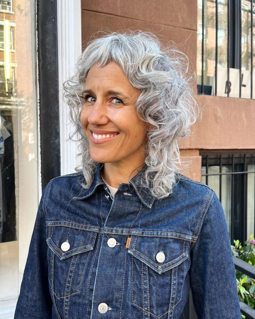 A smiling woman with curly silver hair in a shaggy cut featuring very short micro bangs.