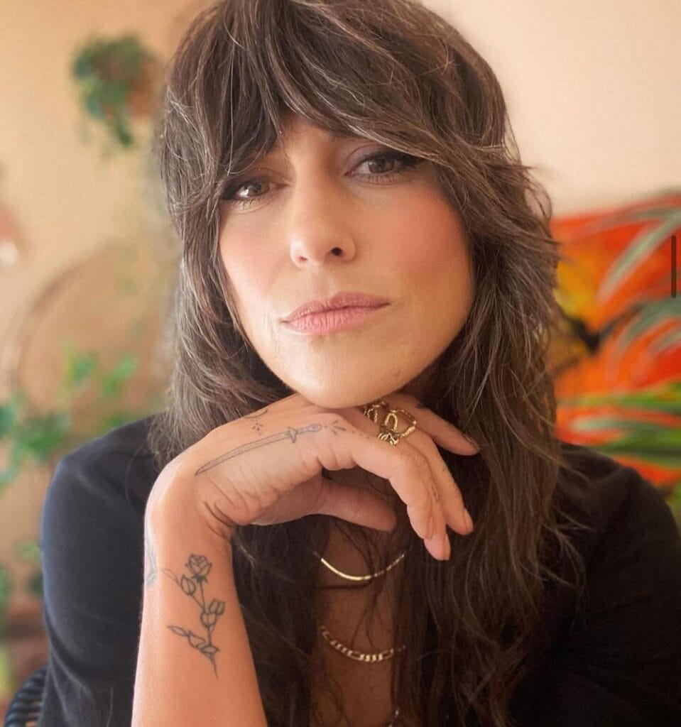 A woman with a long, dark brunette shag featuring heavy curtain bangs and subtle silver highlights.