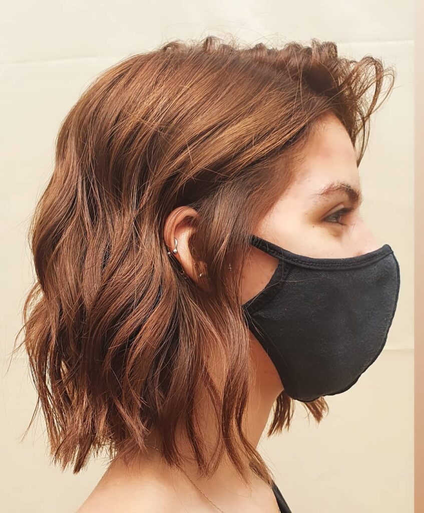 A woman is seen from the side wearing a black mask with a shoulder-length wavy bob in a bright copper brown shade.