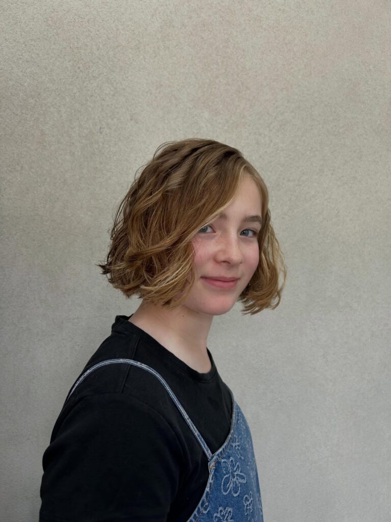A young girl with a chin-length wavy bob in a warm honey-blonde shade featuring a deep side part and tousled texture.