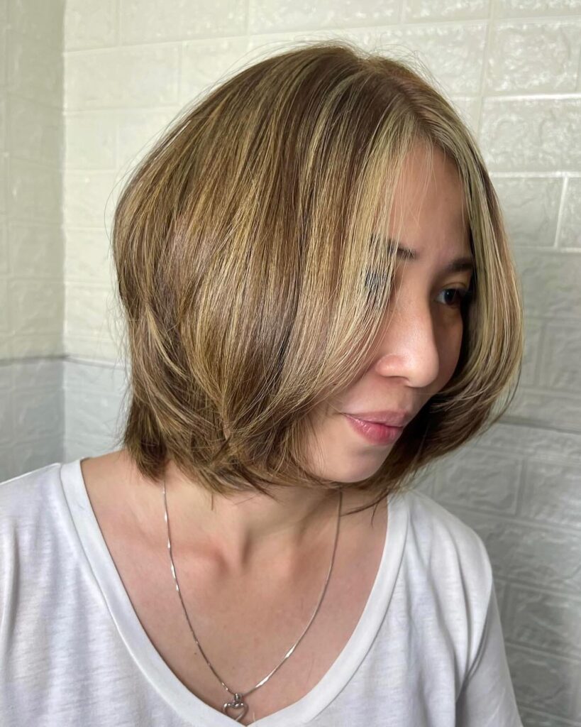 A woman with a rounded chin-length bob in honey brown featuring bright blonde highlights around the face and smooth layers.