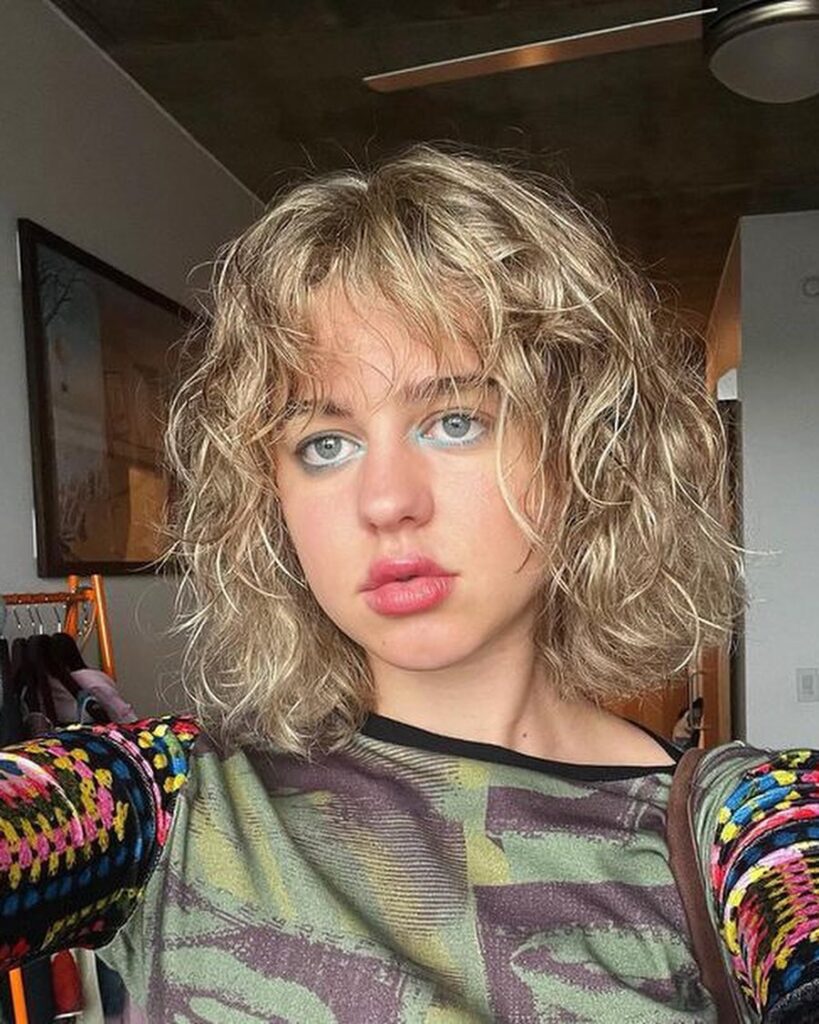 A woman with a voluminous shoulder-length curly bob in dirty blonde, featuring a curly fringe and messy texture.