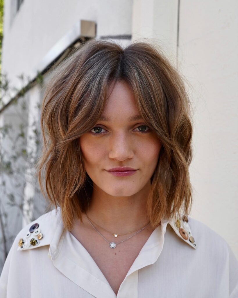 A woman with a shoulder-length wavy lob in sand blonde featuring a middle part and tousled texture.