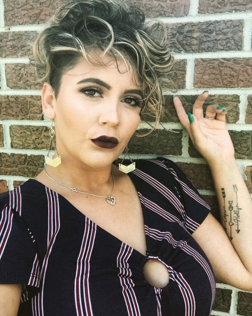 A woman with a curly pixie cut featuring a shaved side and highlighted voluminous curls on top.