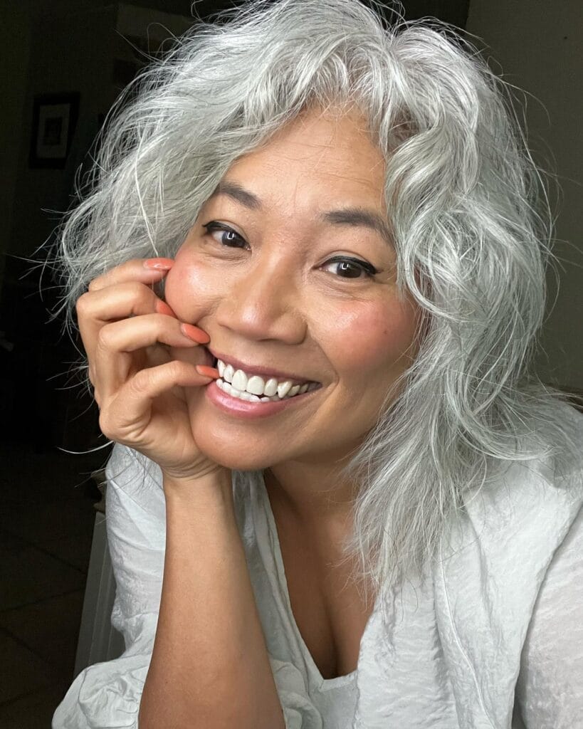 A smiling woman with a shoulder-length silver shaggy hairstyle featuring messy natural waves and soft layers.