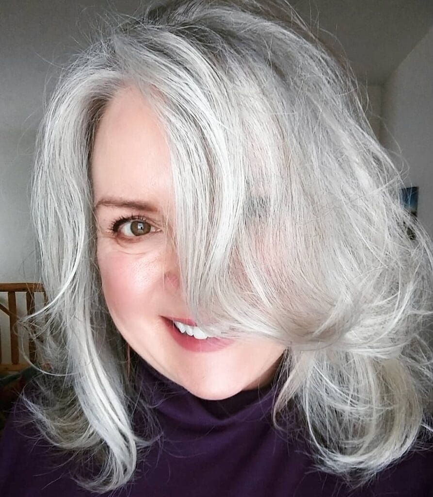 A smiling woman with a voluminous shoulder-length silver hairstyle featuring messy layers and a deep side sweep.