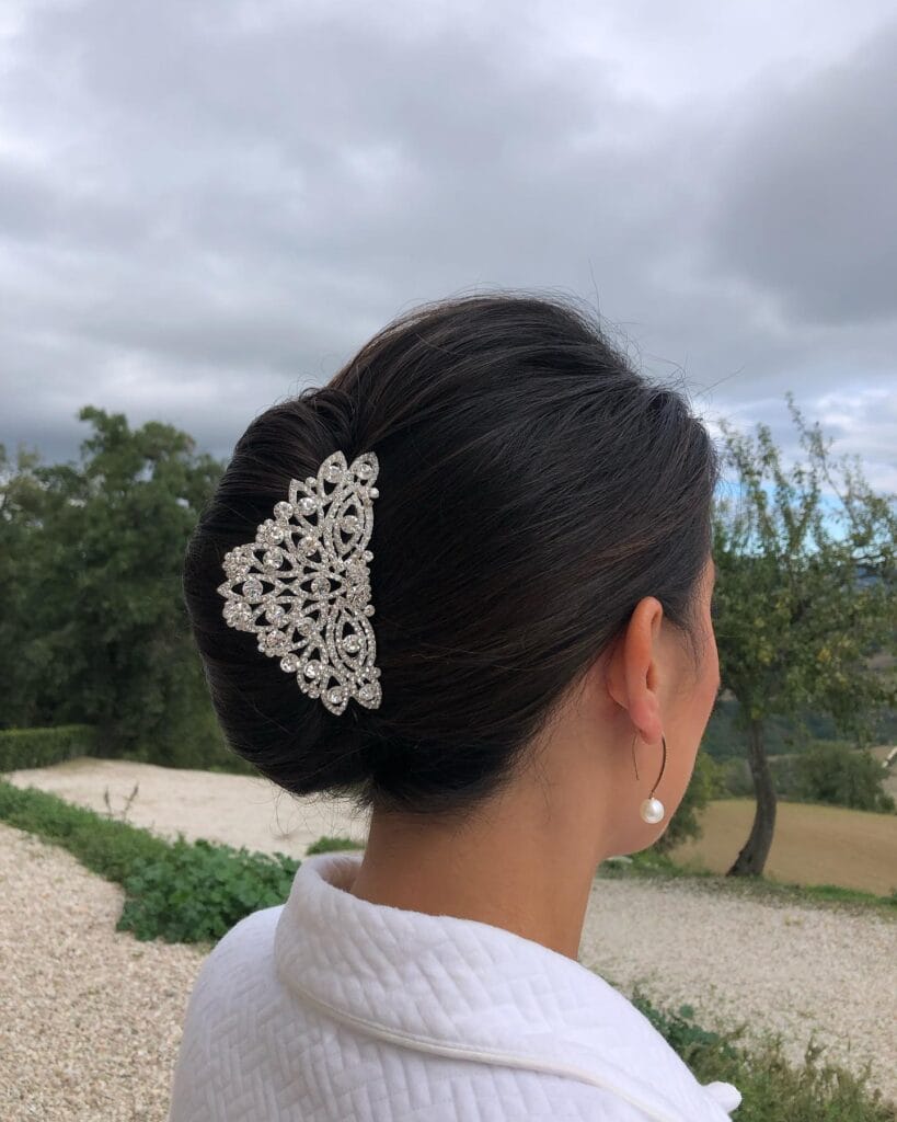 A woman with dark hair viewed from the side and back, styled into a smooth French twist updo, adorned with a large sparkling rhinestone hair clip.