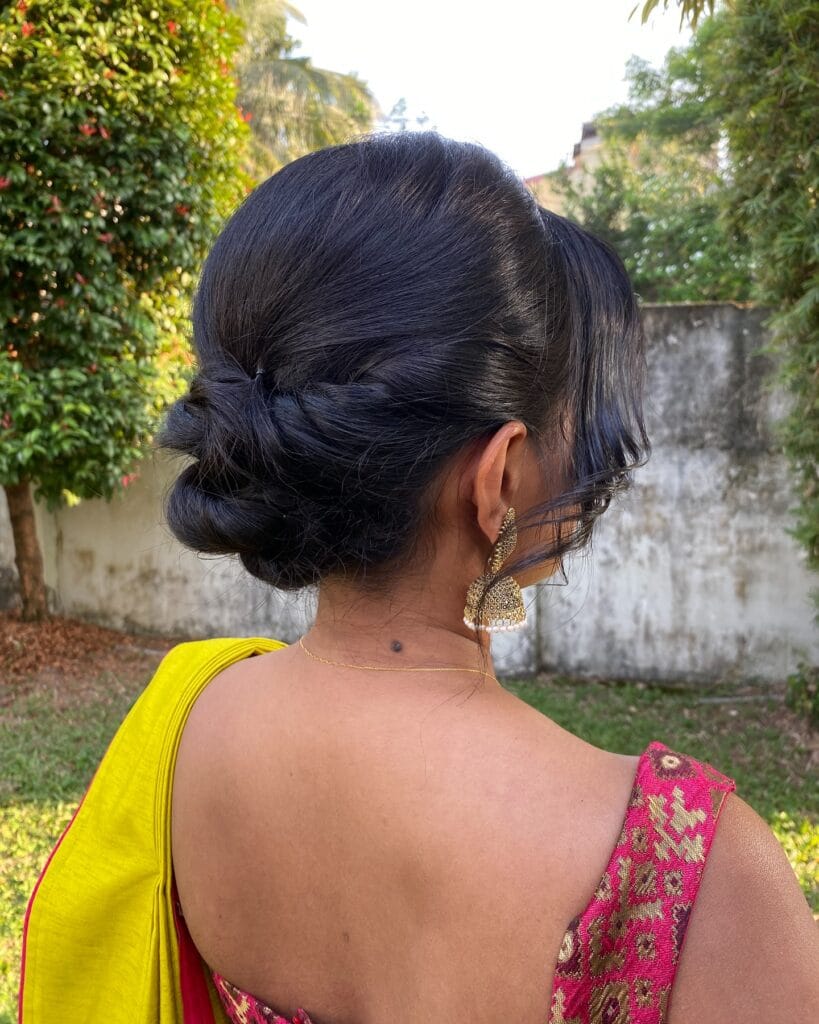 A woman with dark hair is viewed from the side and back, styled into a low twisted bun with soft face-framing curls.