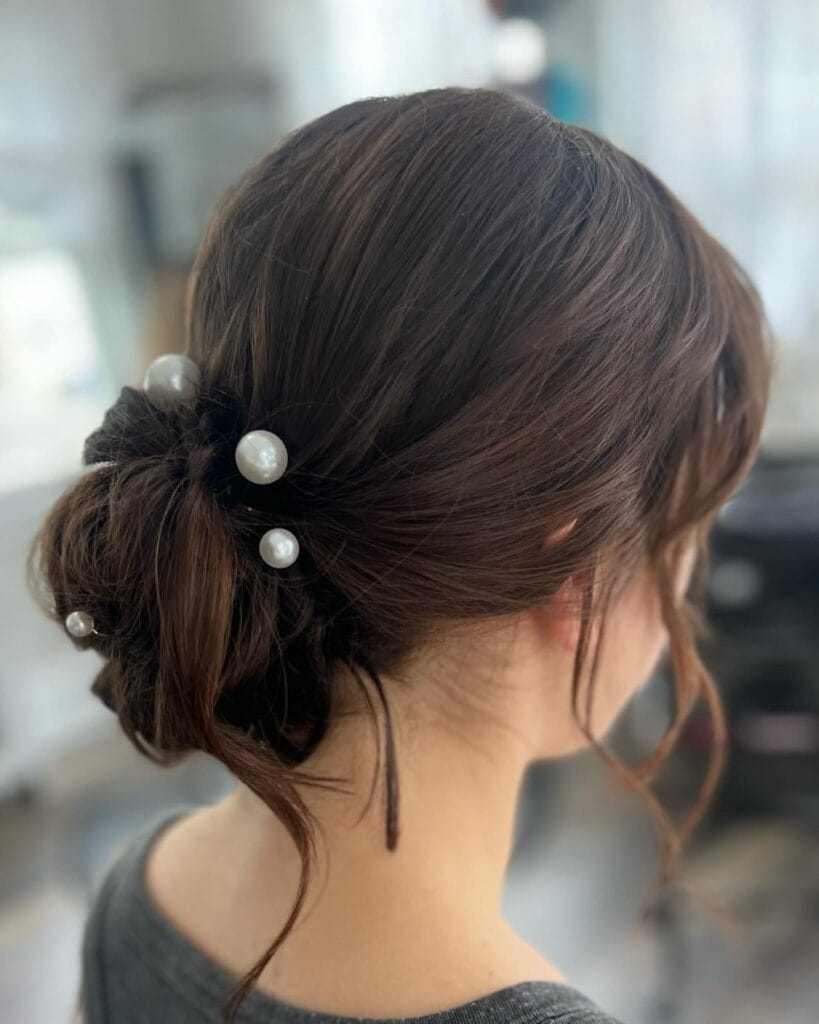 A woman with chestnut brown hair styled in a textured low bun decorated with several white pearl pins.