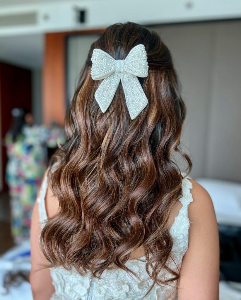 A back view of long, brunette, wavy hair with caramel highlights styled in a half-updo and adorned with a large white pearl and rhinestone bow.