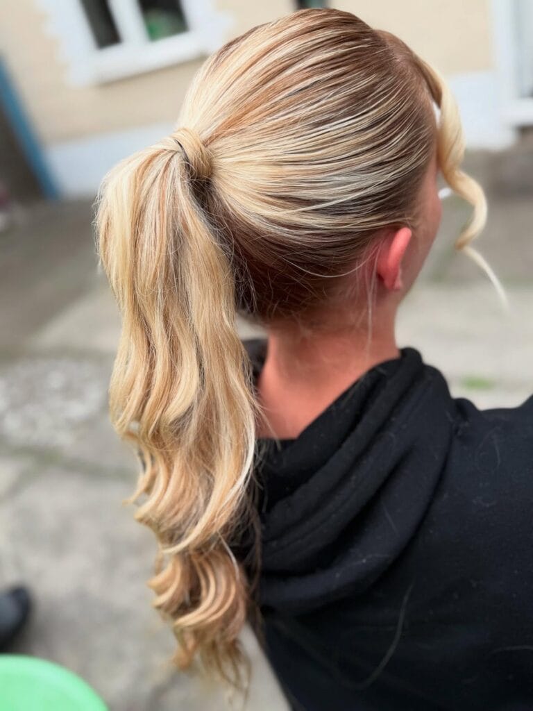 A side and back view of a woman with wavy blonde hair pulled into a medium-height ponytail with a soft face-framing curl.