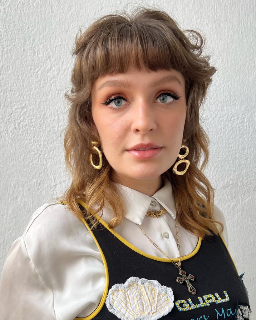 A woman with a shoulder-length honey-blonde wolf cut featuring shaggy layers and blunt straight-across bangs.