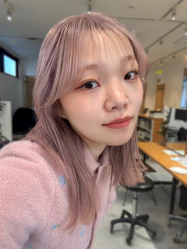 A woman with straight shoulder-length hair in a dusty rose pastel shade featuring thin wispy bangs.