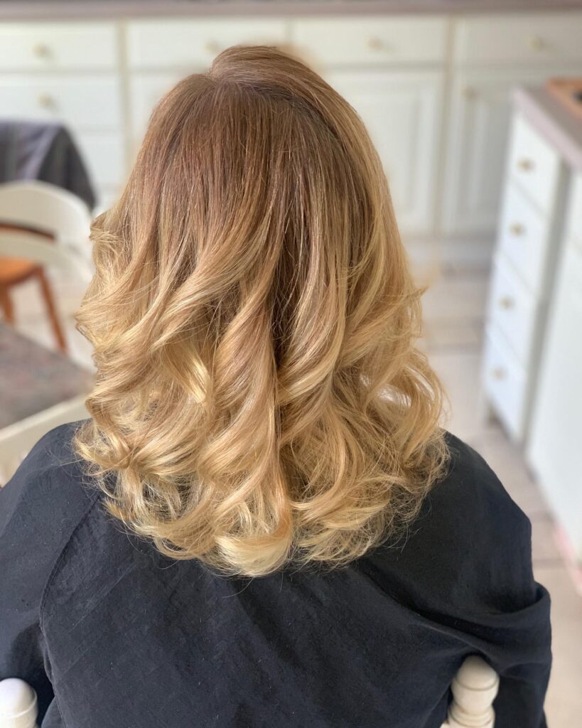 A back view of a woman with shoulder-length, honey blonde hair styled in voluminous, bouncy waves.