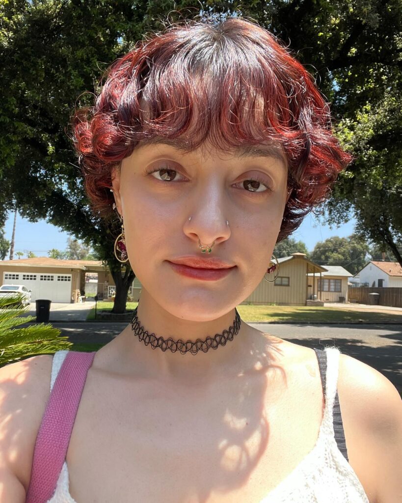 A woman with short wavy hair dyed a vibrant cherry red with wispy bangs and a black choker necklace.