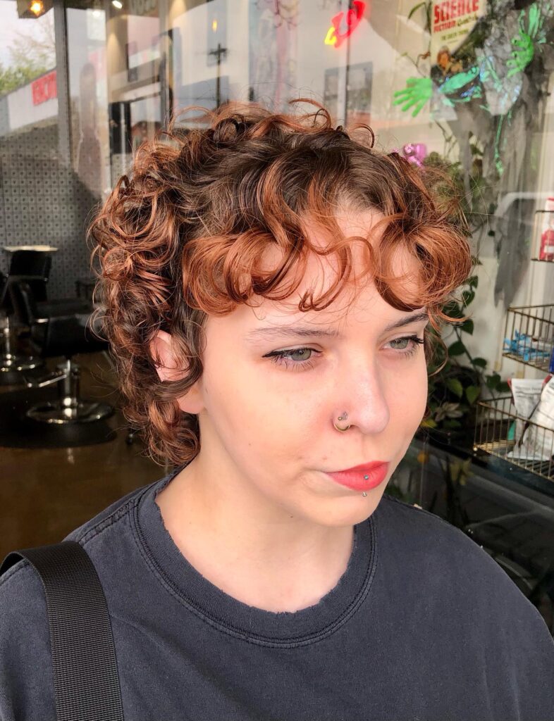 A woman with a short curly pixie cut featuring copper highlights and thick, bouncy bangs.