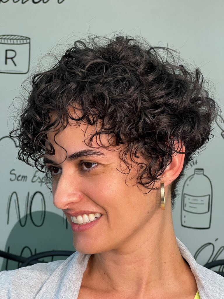 A smiling woman with a short, voluminous, curly pixie cut in a dark brunette shade and curly bangs.